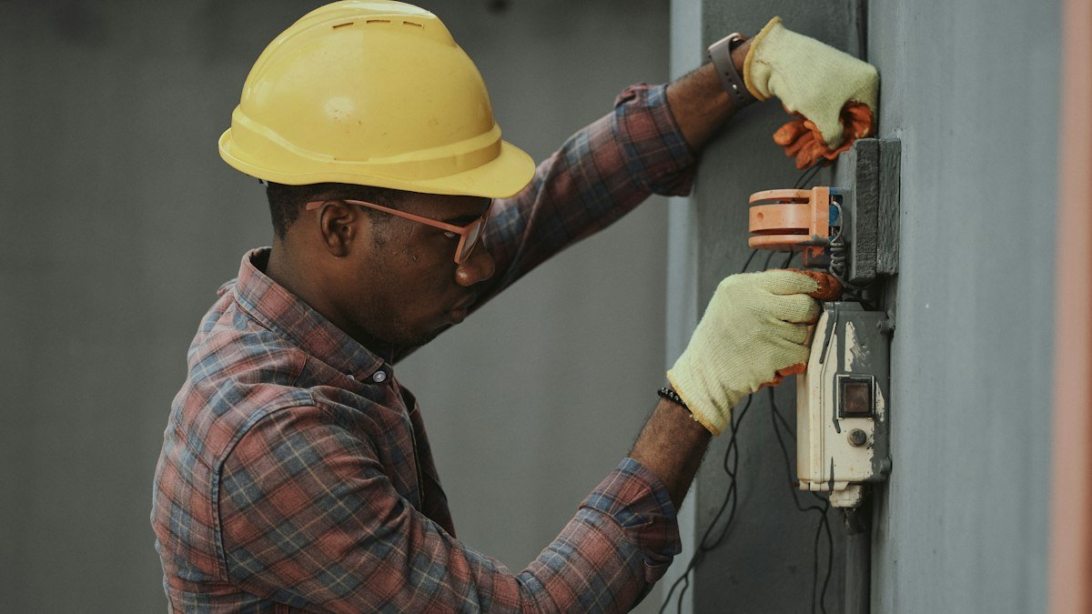 A power worker in a hard hat and plaid shirt tends equipment at a utility site. Photo by Emmanuel Ikwuegbu on Unsplash. Source: https://unsplash.com/photos/man-in-brown-and-white-plaid-dress-shirt-and-yellow-hard-hat-holding-black-and-orange--0-kl1BjvFc (Unsplash License).