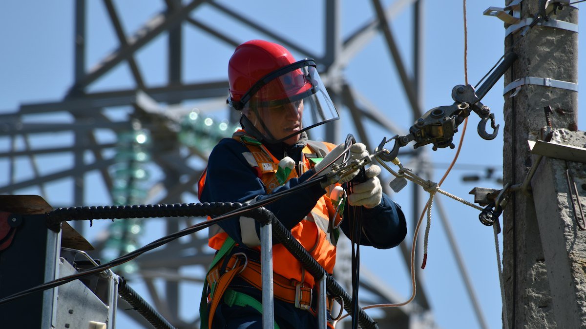A lineworker secures a conductor on a high-voltage transmission tower. Photo by Anton Dmitriev on Unsplash. Source: https://unsplash.com/photos/shallow-focus-photo-of-man-fixing-steel-cable-kBKOaghy8mU (Unsplash License).