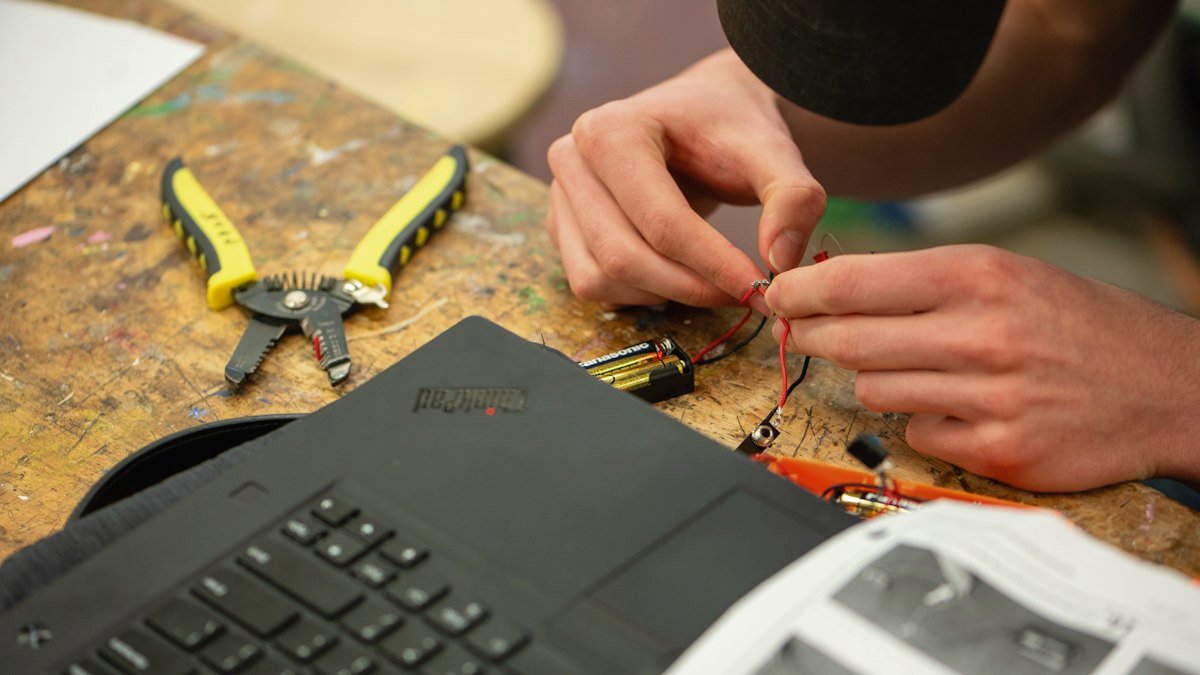 An engineer configures hardware at a workbench with tools and a laptop. Photo by Eric Stoynov on Unsplash. Source: https://unsplash.com/photos/hands-wiring-electronics-with-tools-and-laptop-nearby-y25LI38cd9w (Unsplash License).