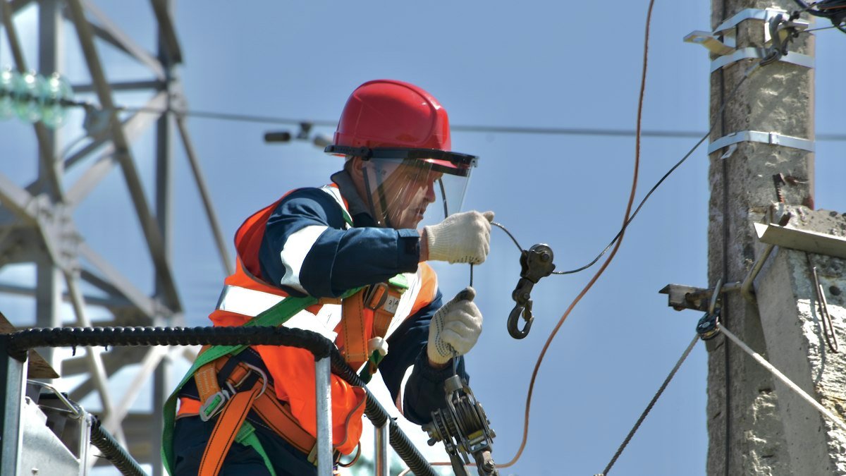 A lineman in safety gear and a red helmet works on a power pole and its lines, standing in for the grid constraints that set the week's data center limits. Photo by Антон Дмитриев on Unsplash. Source: https://unsplash.com/photos/a-man-in-safety-gear-working-on-a-power-pole-ZGY5Pp8Xxaw (Unsplash License).