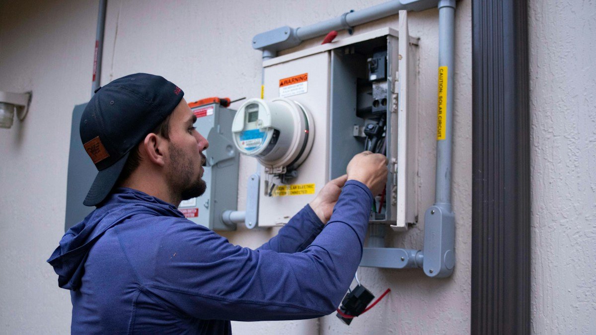 A technician in a cap and blue hoodie working inside an open electrical meter panel. Photo by Raze Solar on Unsplash. Source: https://unsplash.com/photos/person-working-on-electrical-meter-_aSFmmvS62I (Unsplash License).