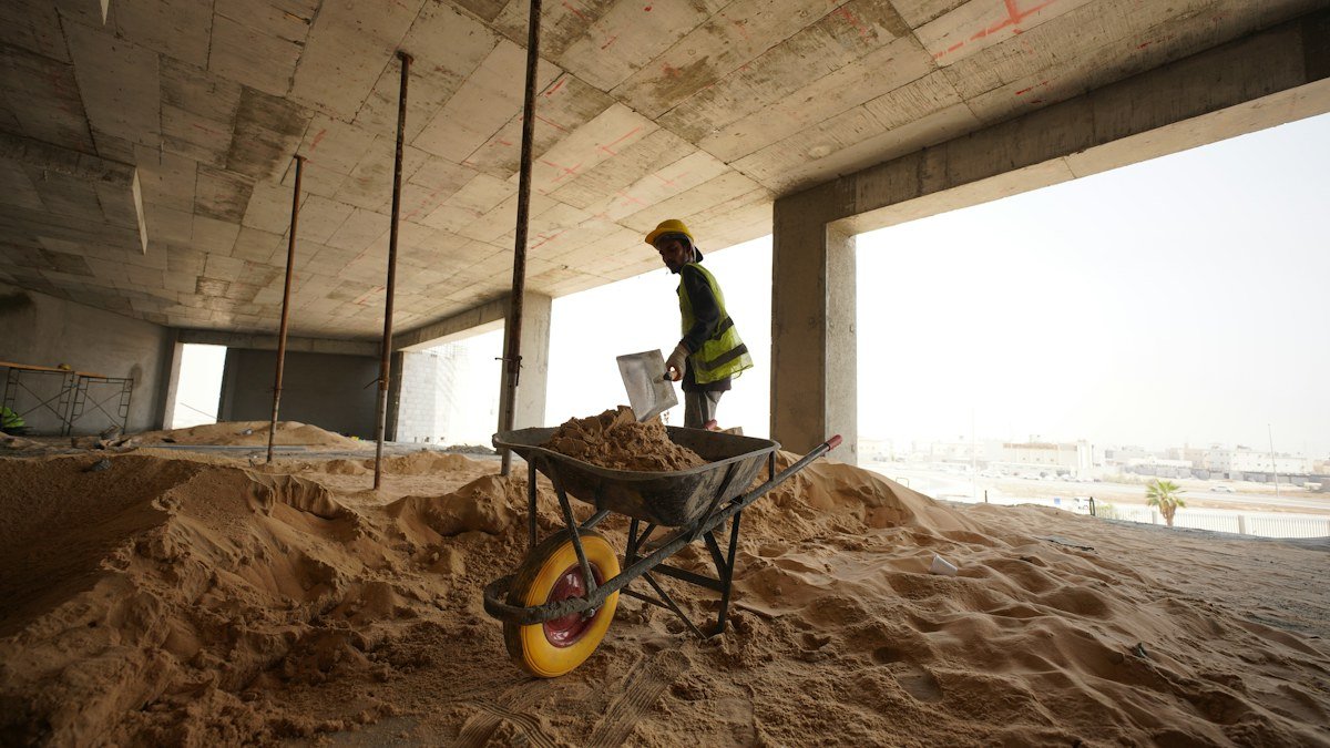 Photo by Glenov Brankovic on Unsplash. Source: https://unsplash.com/photos/a-construction-worker-standing-in-a-pile-of-sand-k3Xa8xH2TPY (Unsplash License). Construction worker in high visibility clothing inside an unfinished building.