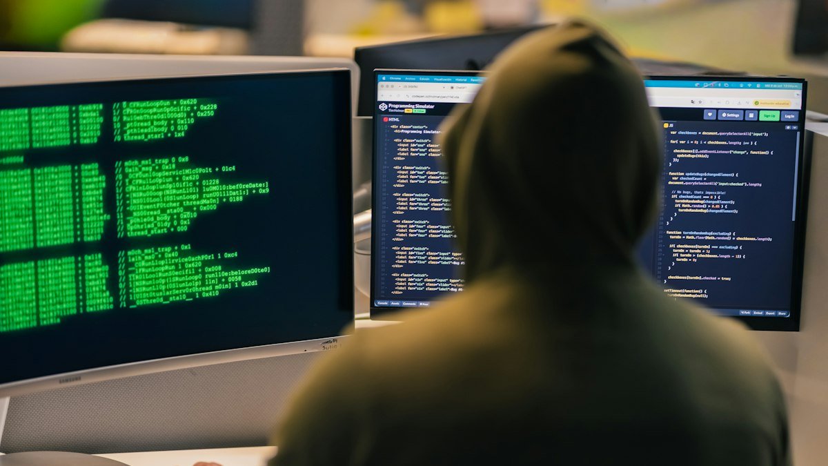 Photo by Julio Lopez on Unsplash. Source: https://unsplash.com/photos/hacker-in-hoodie-working-on-multiple-computer-screens-hLAmfdxGlls (Unsplash License).