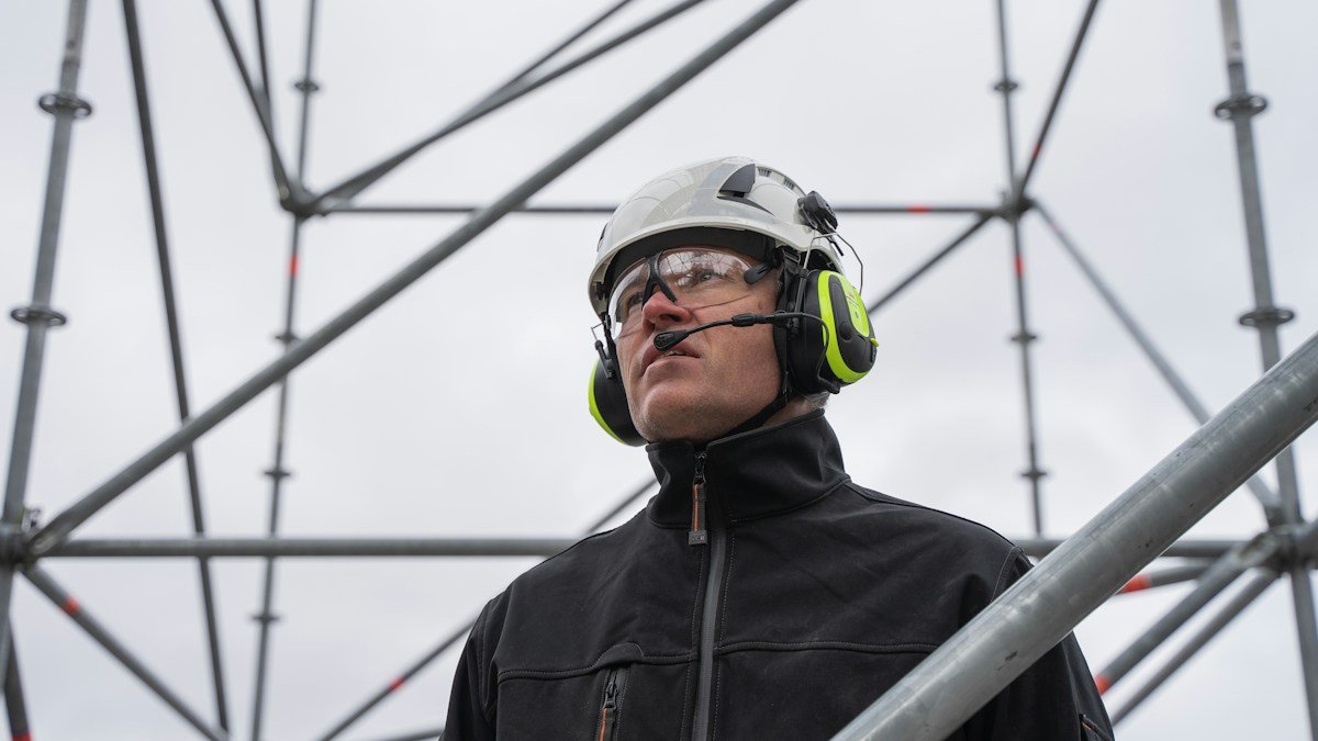 A site worker in a hard hat, safety glasses and ear defenders standing on steel lattice structure at an industrial energy facility. Photo by Jimmy Nilsson Masth on Unsplash (Unsplash License). Source: https://unsplash.com/photos/a-man-wearing-a-helmet-and-goggles-sZdlS32KvNI