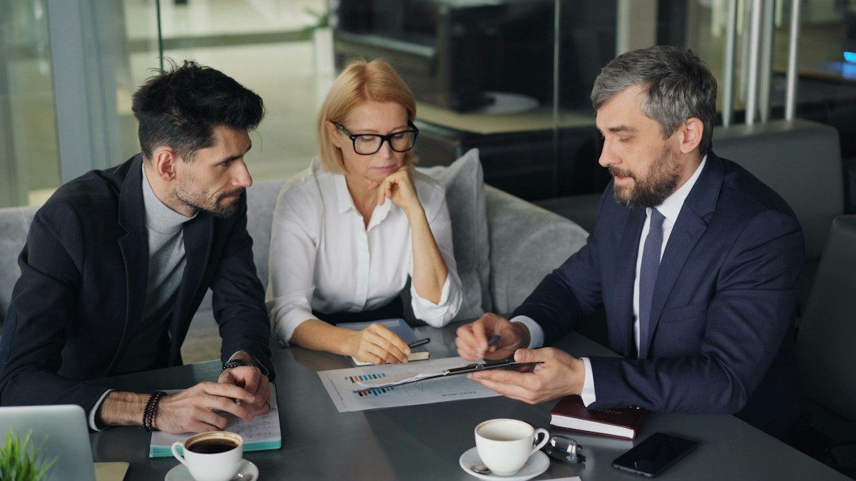 Three colleagues sit around a table reviewing printed deal documents together. Photo by Vitaly Gariev on Unsplash. Source: https://unsplash.com/photos/a-group-of-people-sitting-around-a-table-LS5dCL0NkhE (Unsplash License).