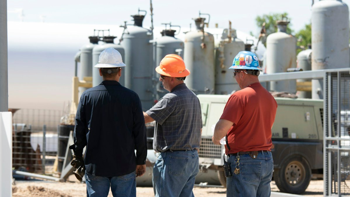 Three workers in hard hats beside industrial process equipment and storage vessels at a power site. Photo by Christina Hawkins on Unsplash (Unsplash License). Source: https://unsplash.com/photos/man-in-black-jacket-standing-beside-man-in-orange-shirt-VDpYOvZm2Ok