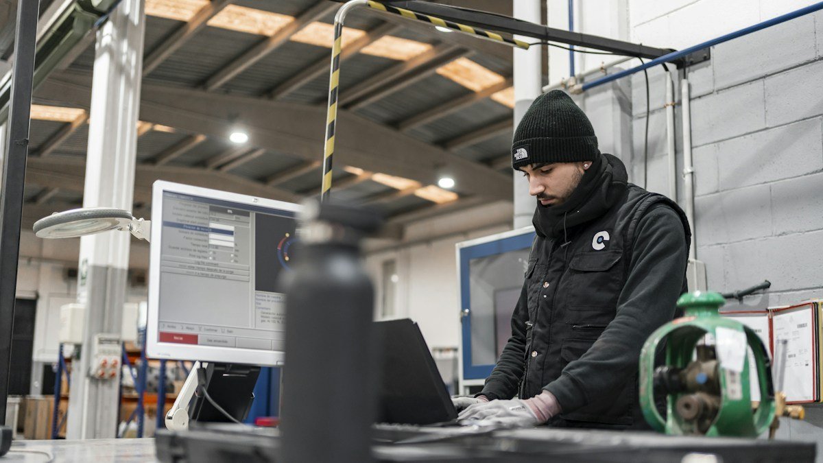 An operator in work gear reads a monitoring screen at a control terminal inside an industrial facility. Photo by TECNIC Bioprocess Solutions on Unsplash. Source: https://unsplash.com/photos/man-works-on-a-computer-in-a-manufacturing-setting-RyMTGAYZpjY (Unsplash License).
